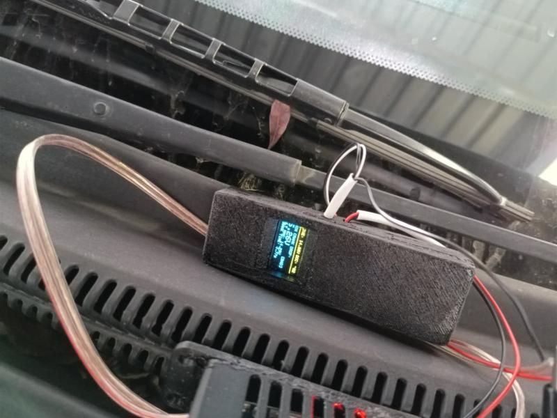 The black 3D-printed monitor box with its lit OLED, resting below a car windscreen with test leads running to the battery