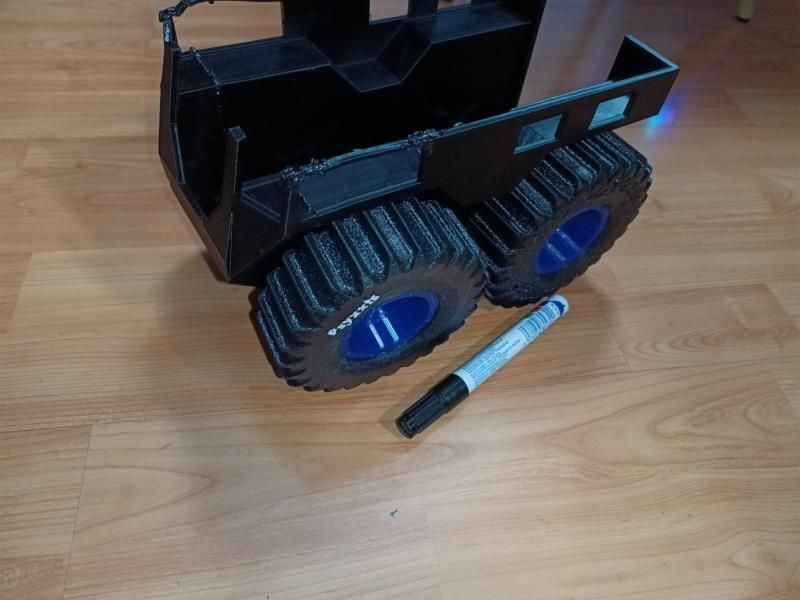 The black 3D-printed crawler body sitting on four chunky black tyres with blue rims, with a marker pen beside it for scale